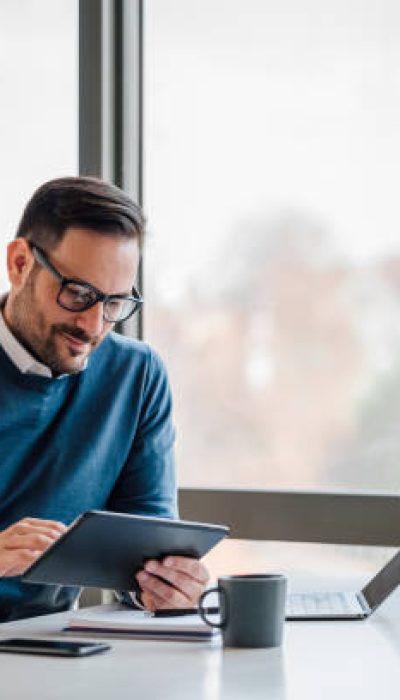 Young businessman using digital tablet while working on laptop in business office. Male professional with wireless computer at desk. He is sitting by window in office.