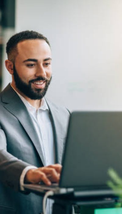 Multiracial Portuguese Jamaican mid adult businessman with beard standing smiling at desk with laptop reviewing data in bright business office wearing suit