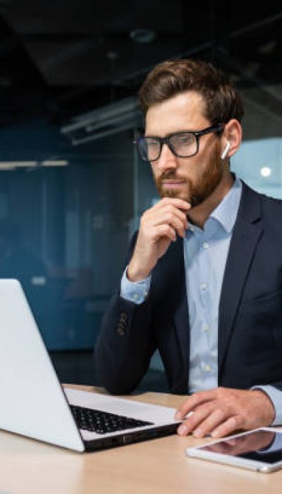 Senior businessman in glasses and beard is thinking about a decision, man is working inside the office and using laptop at work, mature investor in a business suit is sitting at the table